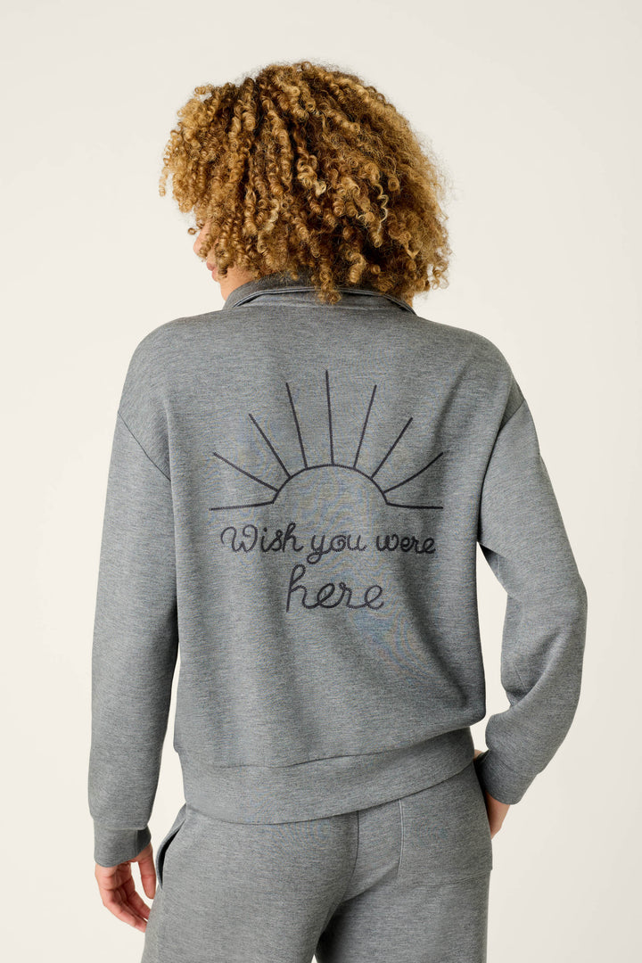 A person with curly hair is wearing the P.J. Salvage “Nature Is My Therapy Wish You Were Here Over Night Pullover” in gray, paired with matching pants. The back of the pullover features a sun illustration and the phrase “Wish you were here.” The background is plain and light.