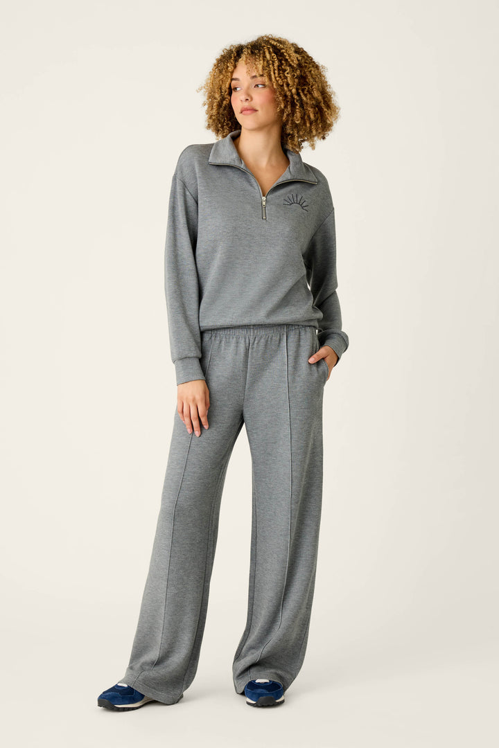 Wearing the P.J. Salvage Nature Is My Therapy Wish You Were Here Over Night Pullover, a person with curly hair stands against a plain light background with one hand in their pocket, pairing the gray zip-up top and matching wide-leg pants with navy sneakers.