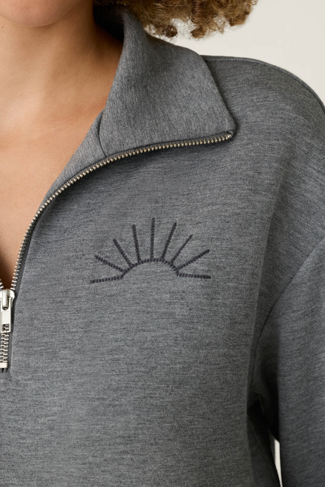 A person wearing the P.J. Salvage Nature Is My Therapy Wish You Were Here Over Night Pullover, a gray zip-up sweater featuring a simple embroidered sunburst design on the left chest.
