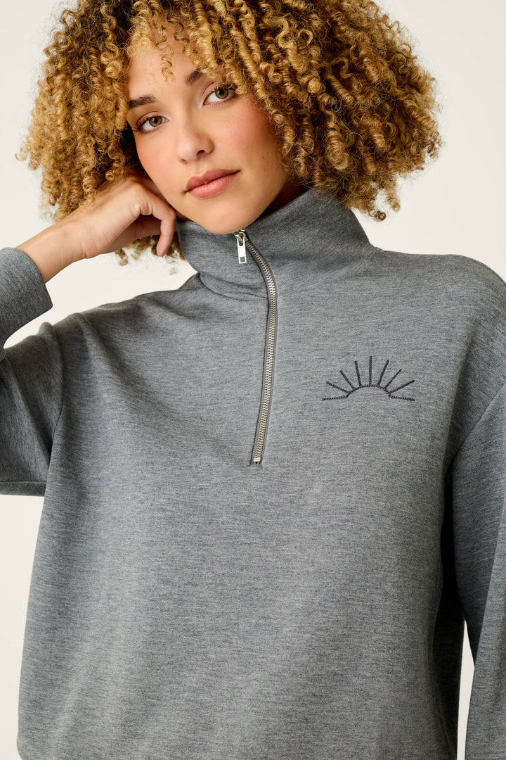 A person with curly hair wears the P.J. Salvage “Nature Is My Therapy Wish You Were Here Over Night Pullover,” a grey quarter-zip sweater featuring a simple embroidered sunburst design on the left side of the chest.