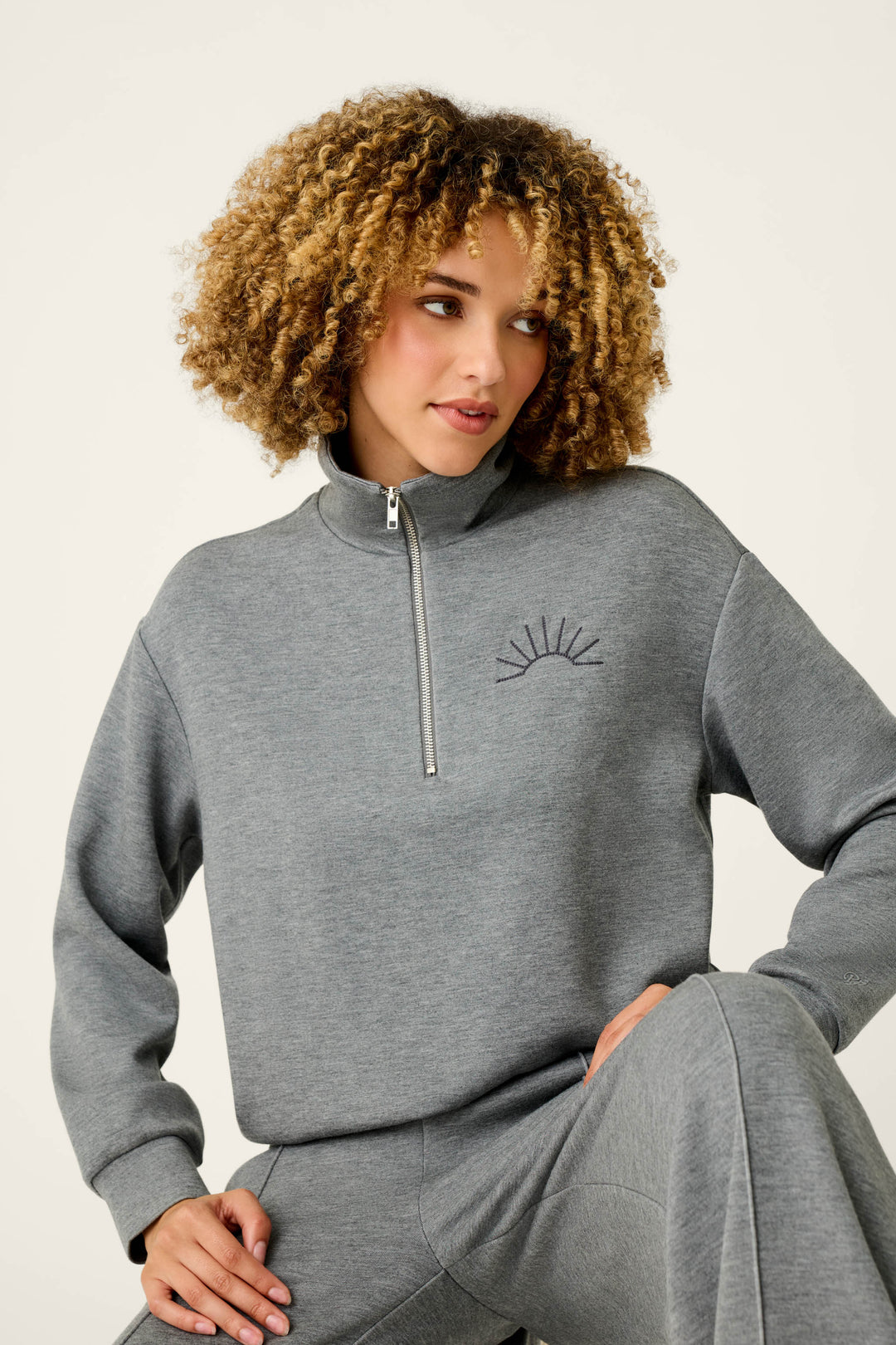 A woman with curly hair is wearing the P.J. Salvage Nature Is My Therapy Wish You Were Here Over Night Pullover in light grey, paired with matching pants. The pullover features a small sunburst graphic on the chest. She is seated and looking to the side, with one hand resting on her knee.