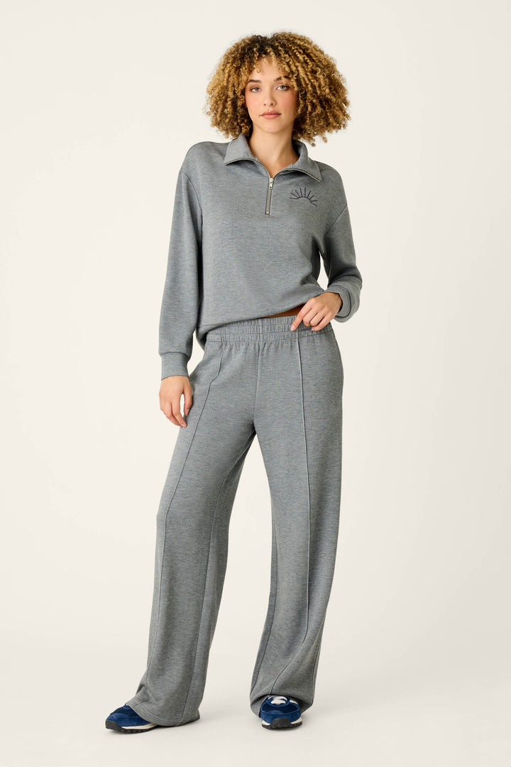 Wearing the Nature Is My Therapy Runway Lounge Pants by P.J. Salvage, a person with curly hair stands against a plain, light background with one hand in their pocket, styled in a matching gray quarter-zip sweater and wide-leg pants.