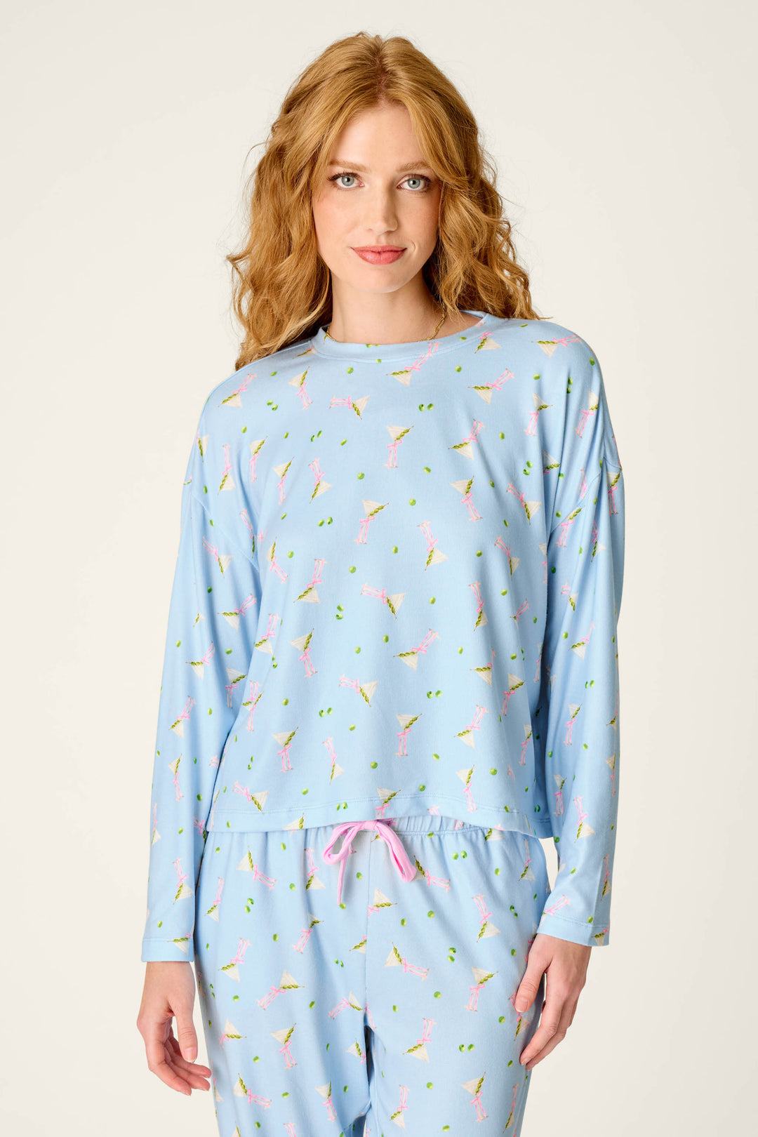 A woman with wavy red hair is wearing the P.J. Salvage Olive You Martini Print Long Sleeve Top, featuring a light blue fabric patterned with cocktail glasses and accented by a pink drawstring, standing in front of a plain background.