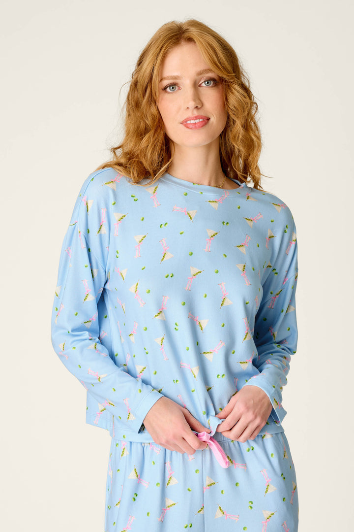 A woman with wavy red hair wears the P.J. Salvage Olive You Martini Print Long Sleeve Top. She stands against a plain background, smiling gently as she ties a knot in her top.