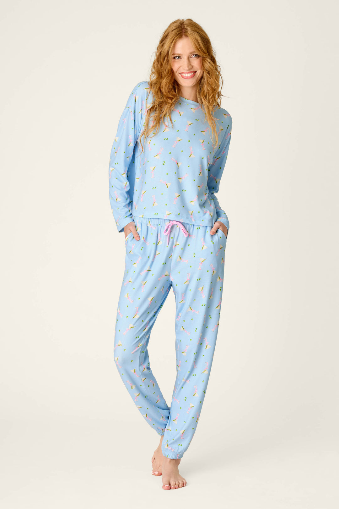 A woman with long, wavy hair smiles while wearing the P.J. Salvage Olive You Martini Print Long Sleeve Top in light blue with a colorful martini print. She stands barefoot, one leg crossed in front of the other, hands in her pants pockets against a plain background.