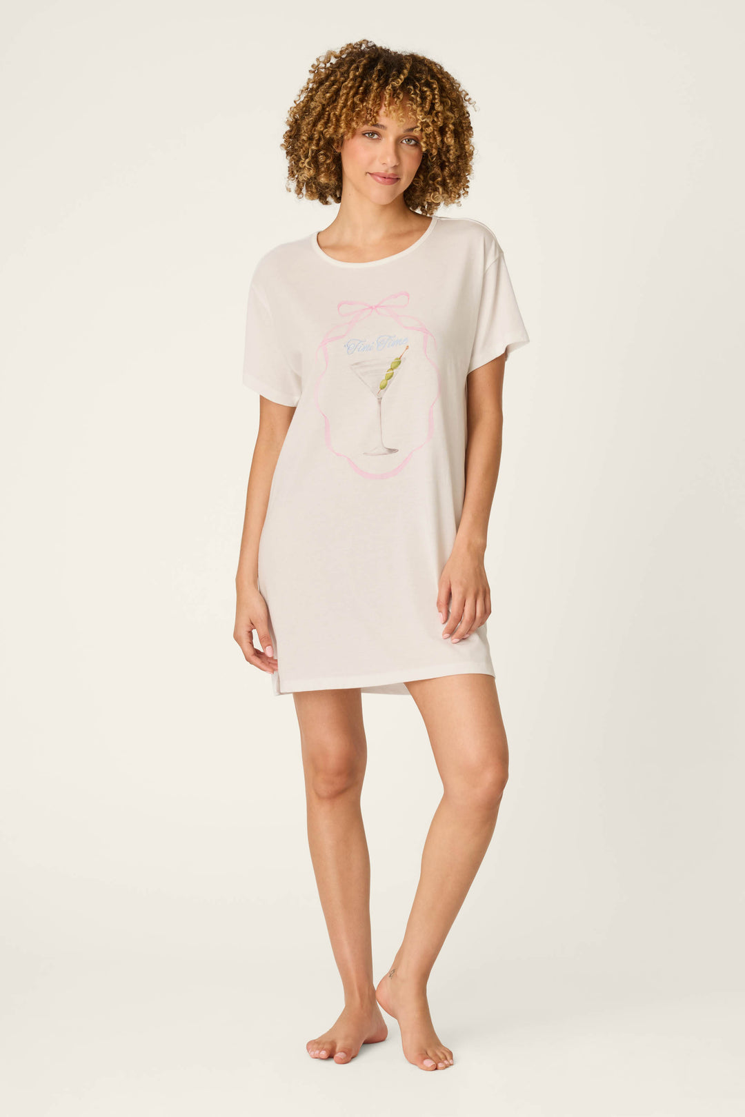 A person with curly hair wears the P.J. Salvage Olive You Martini Print Nightshirt, a loose, short-sleeved, light-colored sleep shirt featuring a drink graphic on the front, standing barefoot against a plain background.
