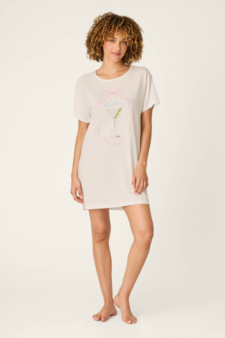 A person with curly hair wears the P.J. Salvage Olive You Martini Print Nightshirt, a loose, short-sleeved, light-colored sleep shirt featuring a drink graphic on the front, standing barefoot against a plain background.