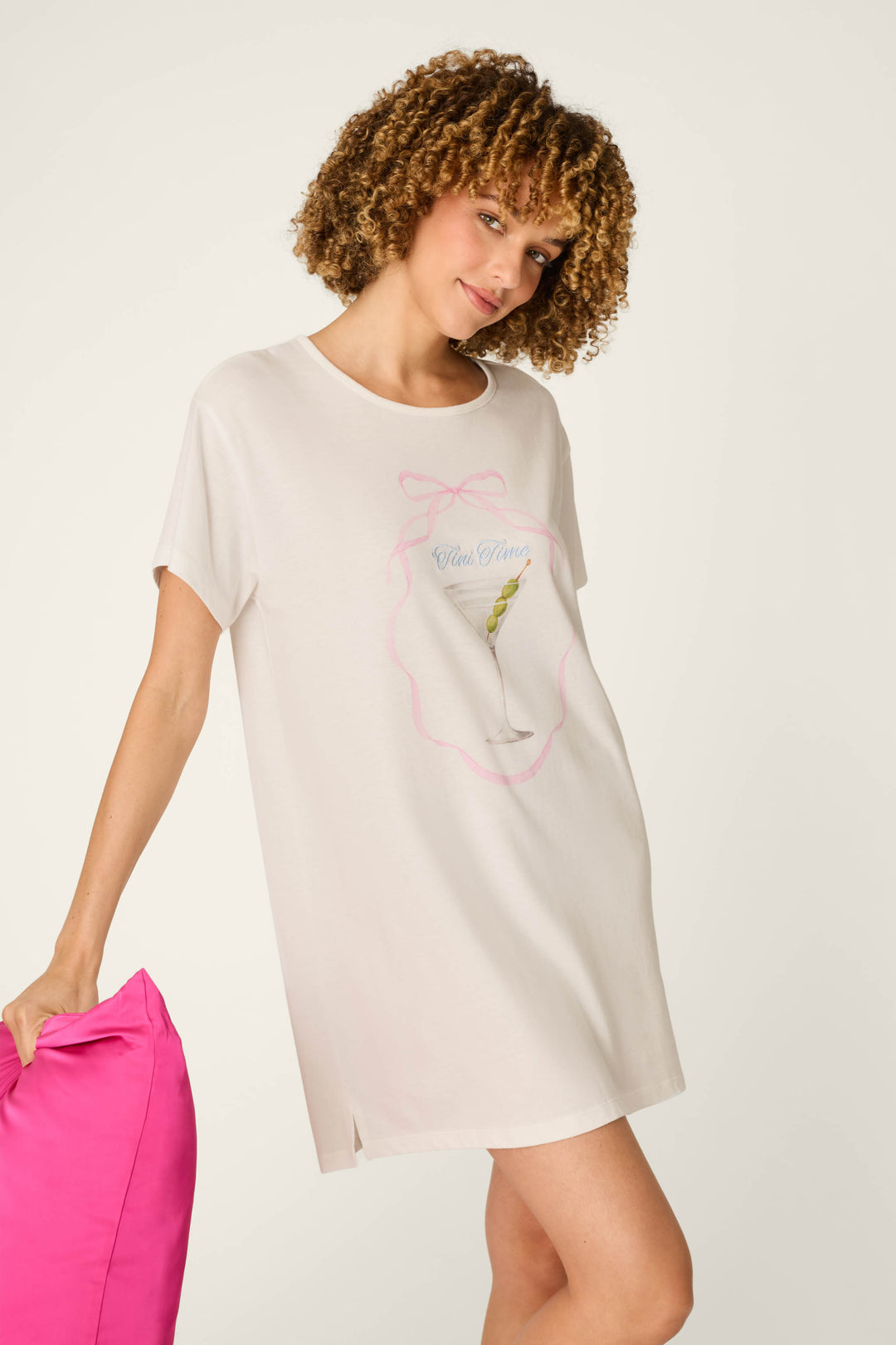 A woman with curly hair wears the Olive You Martini Print Nightshirt by P.J. Salvage—a short-sleeve, white T-shirt dress featuring a martini glass graphic—while holding a bright pink pillow, standing against a plain light background.