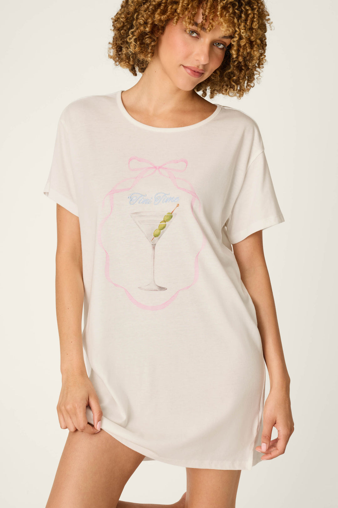 A woman wearing the P.J. Salvage Olive You Martini Print Nightshirt, which features a martini glass with olives and playful text, stands against a light background with a slight smile.