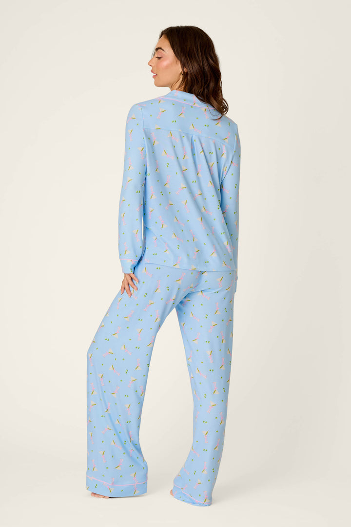 A woman stands barefoot, facing away, wearing the P.J. Salvage Olive You Martini Print Long PJ Set—a light blue pajama set with a small, colorful martini print. The outfit features long pants and a long-sleeve top against a plain off-white background.