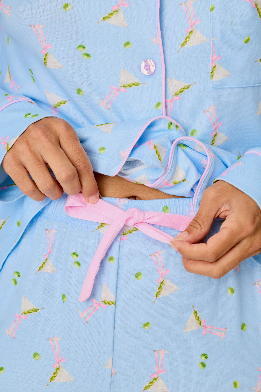 A person, visible only from the hands to midsection, ties a pink drawstring on pajama pants while wearing the Olive You Martini Print Long PJ Set by P.J. Salvage. The pajamas feature green and pink martini glass designs with white trim on a blue patterned background.
