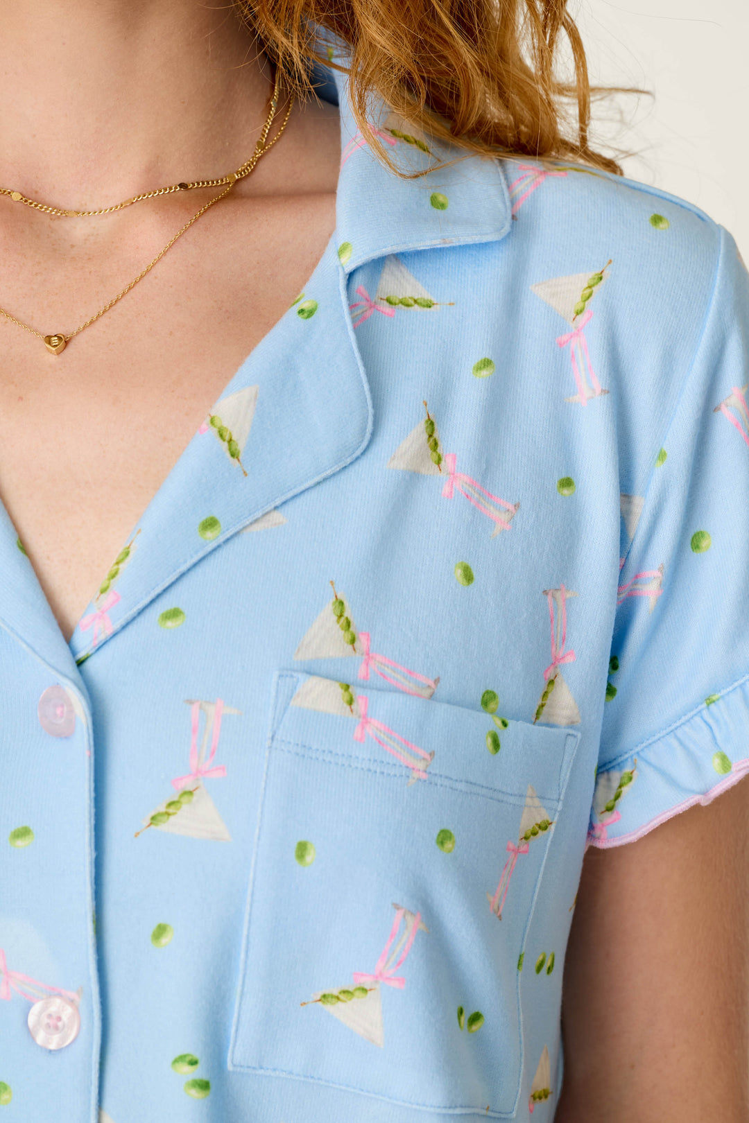 A person is wearing the P.J. Salvage Olive You Martini Print Short PJ Set, which features a blue shirt adorned with pink cocktails, green olives, and white triangles, complete with a front pocket and collar. They accessorize with two delicate gold necklaces.