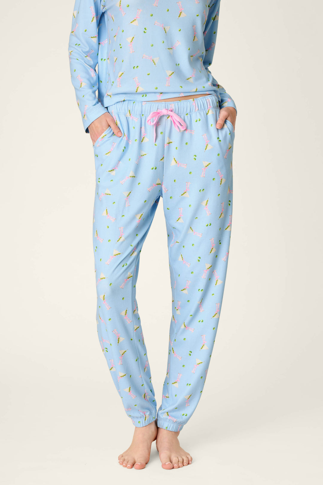 A person is wearing P.J. Salvage Olive You Martini Print Jogger Pajama Pants, featuring a playful martini glass pattern and pink drawstring, standing barefoot on a plain background with their upper body partially visible and hands in their pockets.