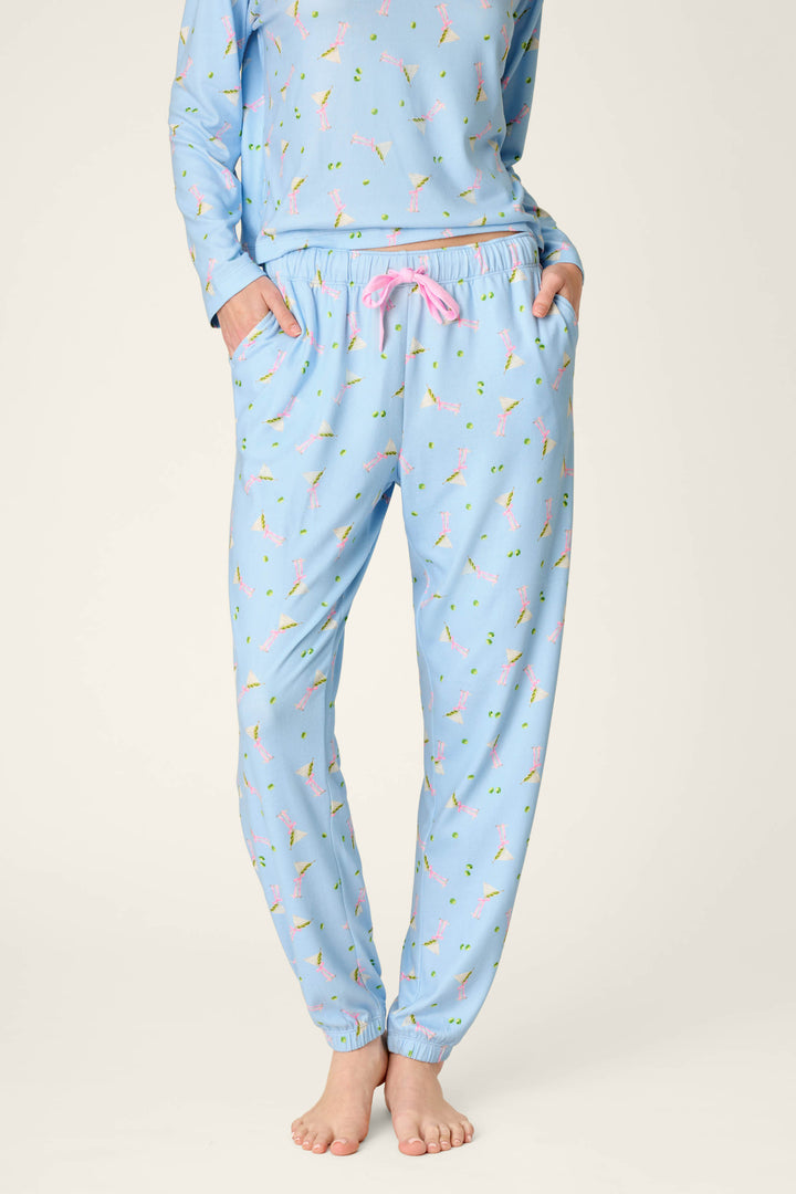 A person is wearing P.J. Salvage Olive You Martini Print Jogger Pajama Pants, featuring a playful martini glass pattern and pink drawstring, standing barefoot on a plain background with their upper body partially visible and hands in their pockets.