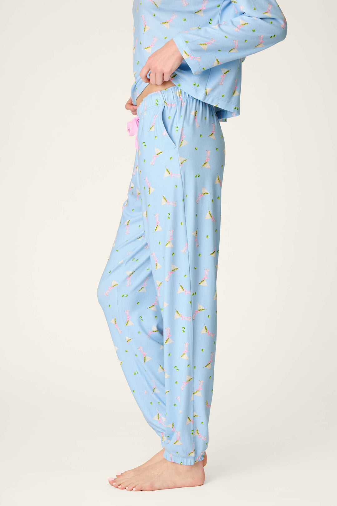 A person is wearing the Olive You Martini Print Jogger Pajama Pants by P.J. Salvage, featuring a colorful, small martini glass pattern on light blue fabric. The side view image captures them from shoulders to bare feet against a plain background.