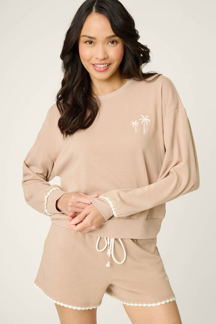 A woman smiles against a light background wearing the P.J. Salvage Palms Away Warm Taupe Sweatshirt, a light beige long-sleeve top with white trim and a small palm tree graphic, paired with matching shorts.