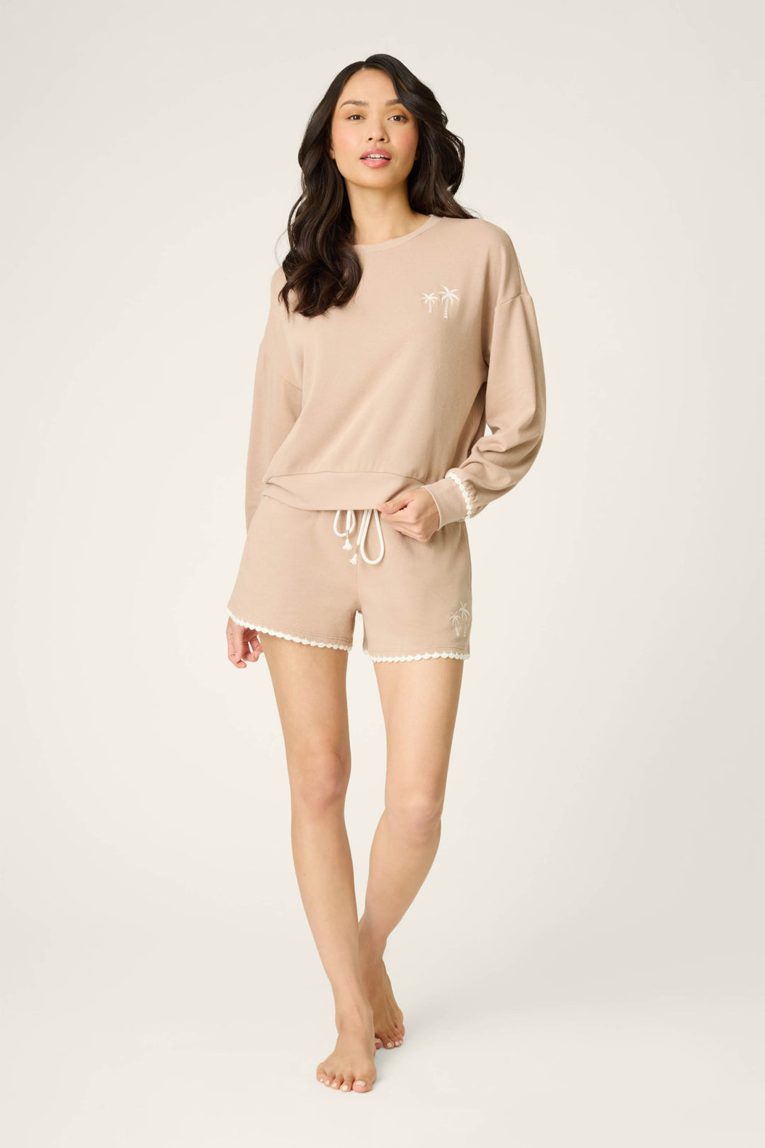 A woman with long dark hair wears the P.J. Salvage Palms Away Warm Taupe Sweatshirt, featuring white palm tree designs, paired with matching drawstring shorts. She stands barefoot against a plain light background.