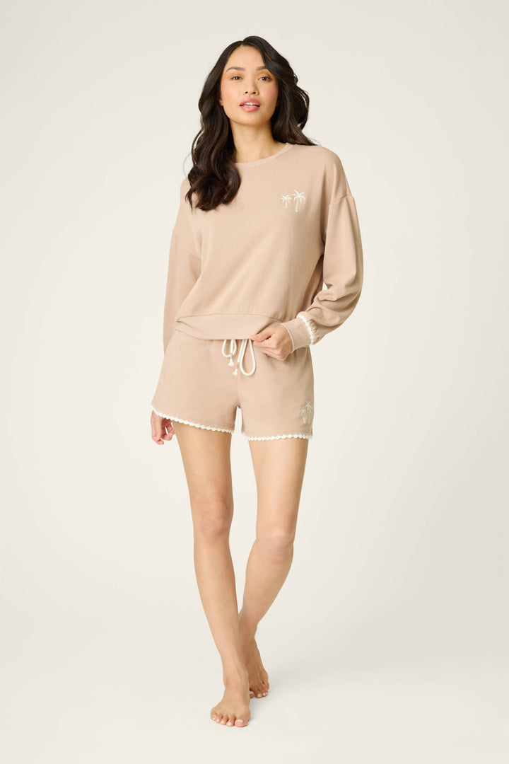 A woman with long dark hair wears the P.J. Salvage Palms Away Warm Taupe Sweatshirt, featuring white palm tree designs, paired with matching drawstring shorts. She stands barefoot against a plain light background.