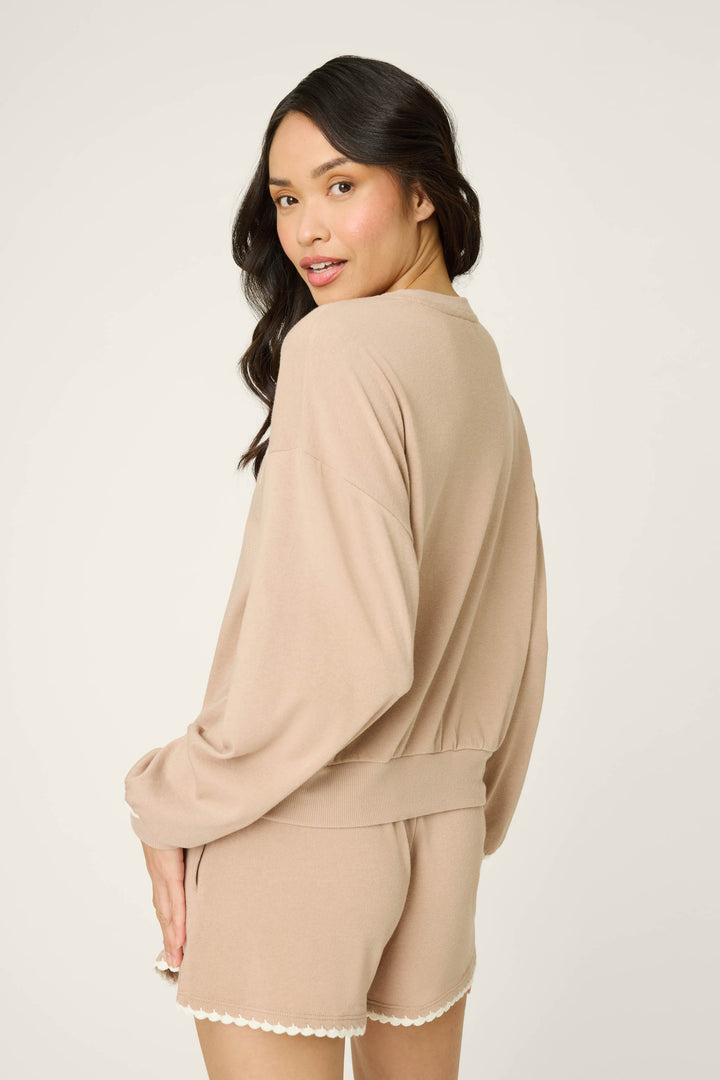 A woman with long, wavy dark hair models the P.J. Salvage Palms Away Warm Taupe Sweatshirt, turning slightly to the side and smiling softly as she looks back over her shoulder.