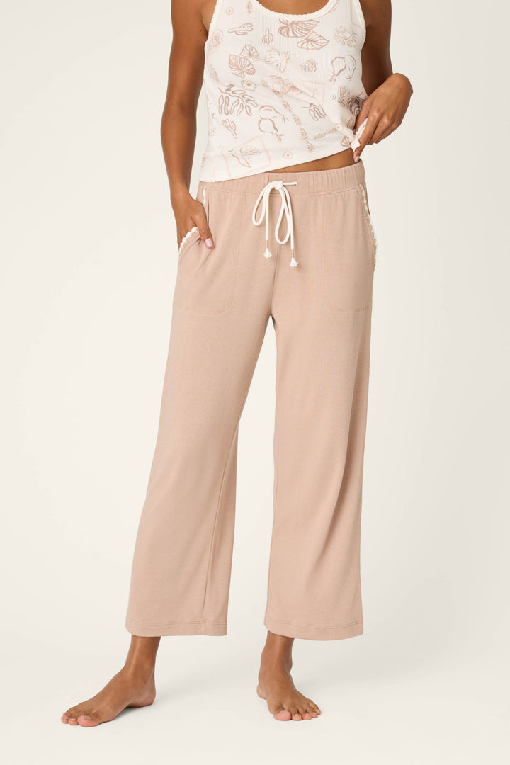 A person stands barefoot in P.J. Salvage’s Palms Away Warm Taupe Open Leg Pants, paired with a sleeveless white top featuring a pale seashell pattern, posing against a neutral background.