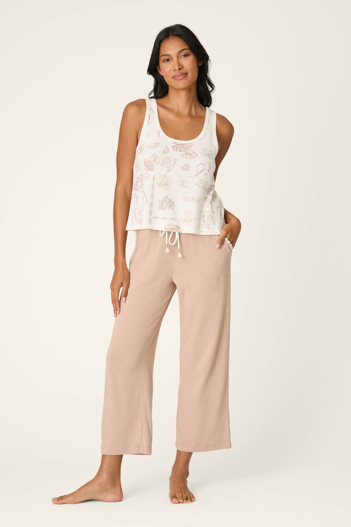 A woman with long dark hair poses barefoot against a light background, wearing a sleeveless white patterned tank top and P.J. Salvage Palms Away Warm Taupe Open Leg Pants, with one hand in her pocket.