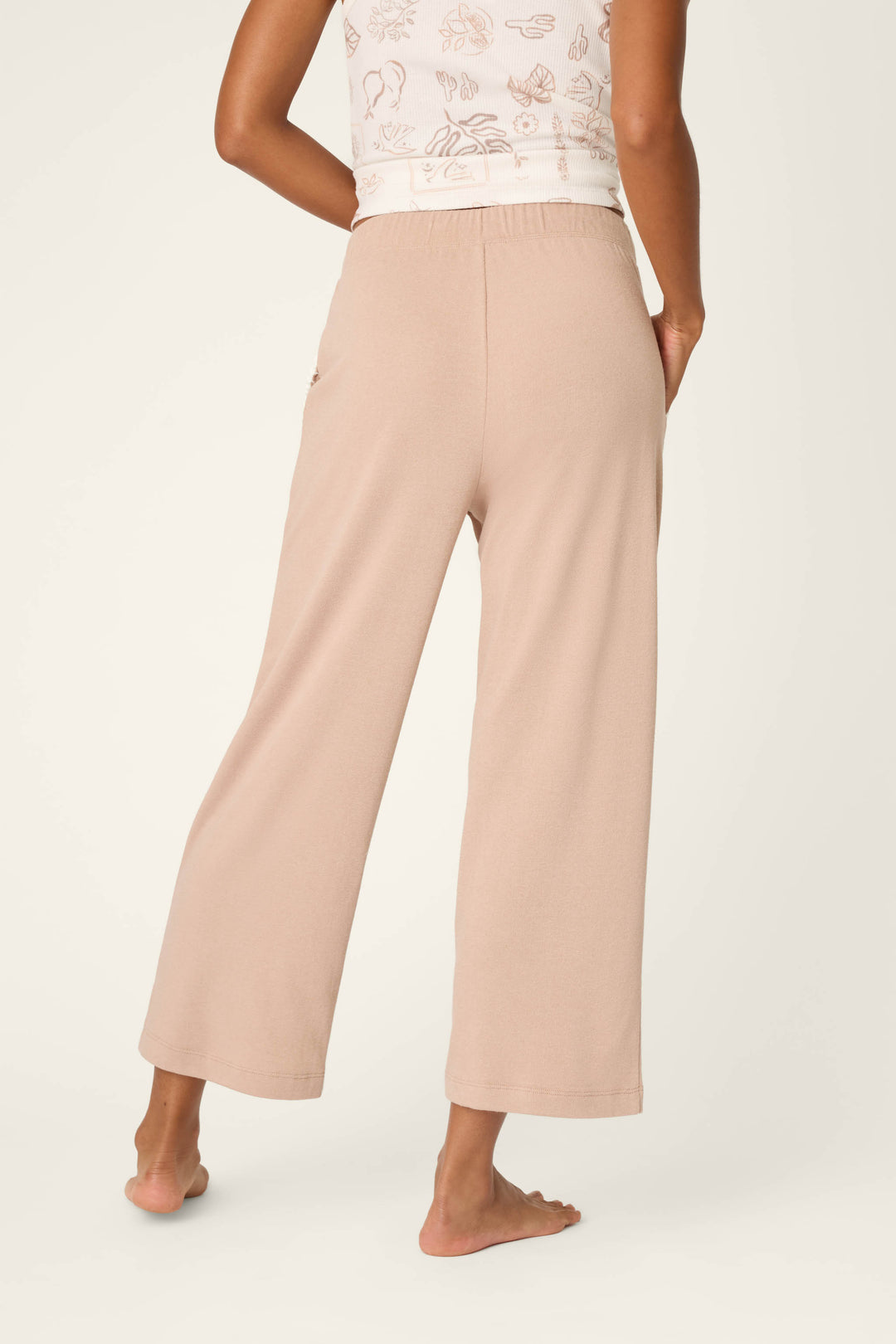 A person stands barefoot facing away, wearing P.J. Salvage Palms Away Warm Taupe Open Leg Pants and a white top with a pale abstract print against a neutral background.