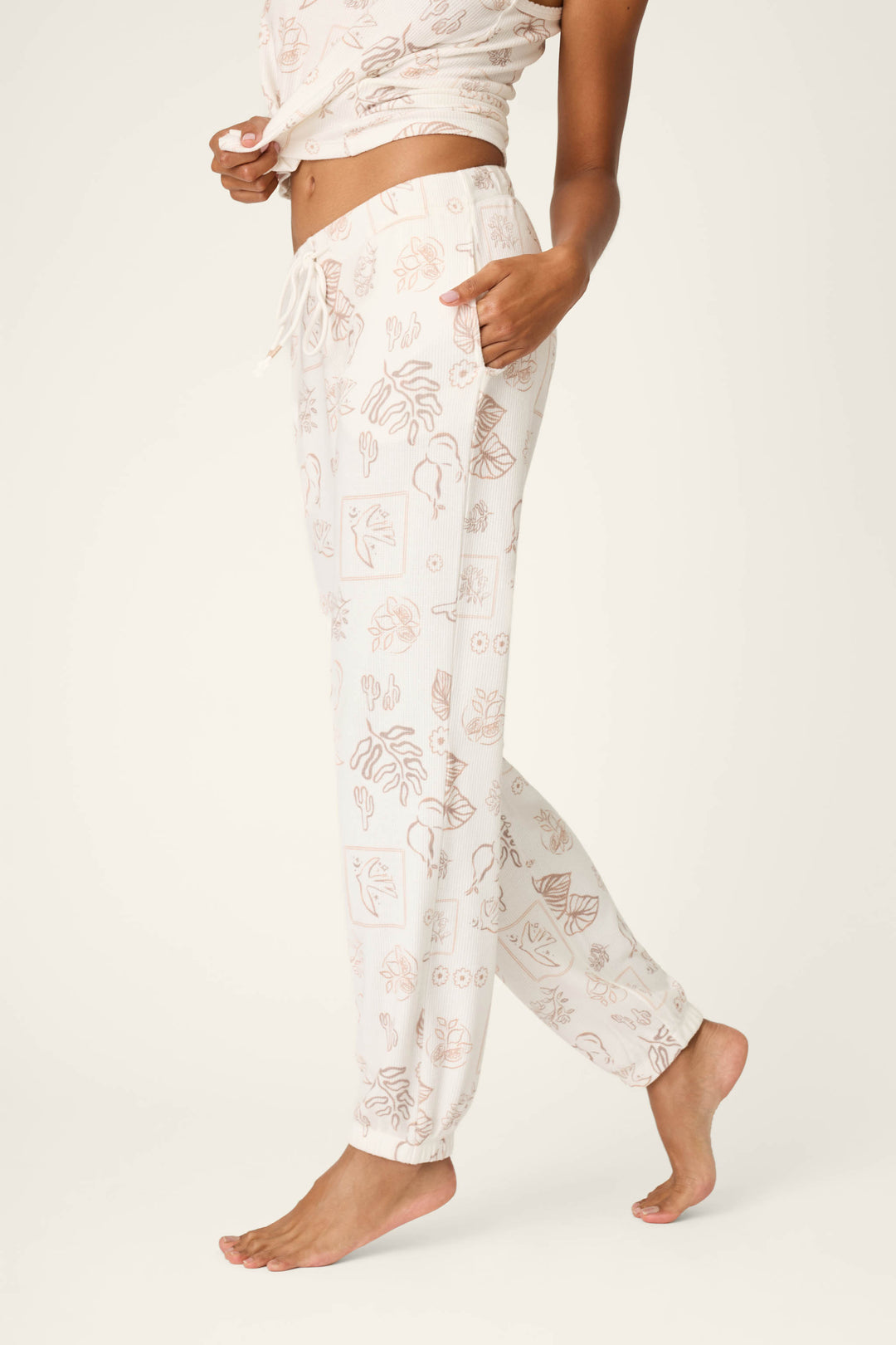 A person stands barefoot in P.J. Salvage Palms Away Bistro Botanicals Banded Pants, featuring a light-colored, subtle botanical and abstract print, paired with a matching top against a plain off-white background.