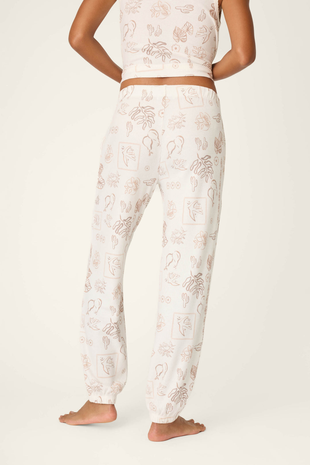A person stands barefoot, shown from the back, wearing P.J. Salvage Palms Away Bistro Botanicals Banded Pants with a matching cropped tank top featuring abstract line art and plant patterns against a plain off-white background.