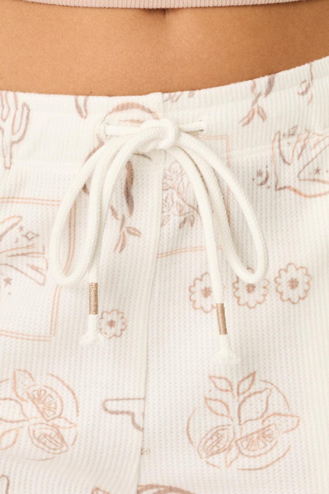 Close-up of a person's midsection wearing P.J. Salvage Palms Away Bistro Botanicals Shorts—white, textured with a drawstring and light brown botanical line art including flowers, citrus slices, and cacti. A beige top's waistband is visible above.