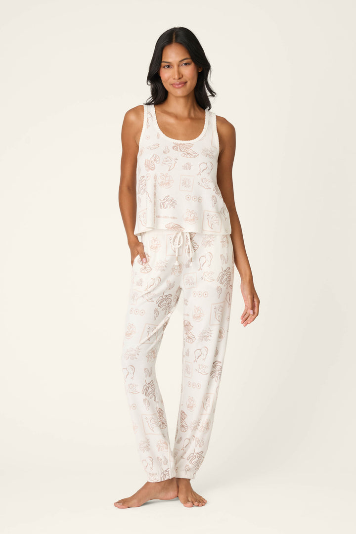 A woman stands barefoot against a plain background, smiling softly with one hand in her pocket, wearing the P.J. Salvage Palms Away Bistro Botanicals Long PJ Set: a white sleeveless pajama set with light brown whimsical patterns and drawstring pants.