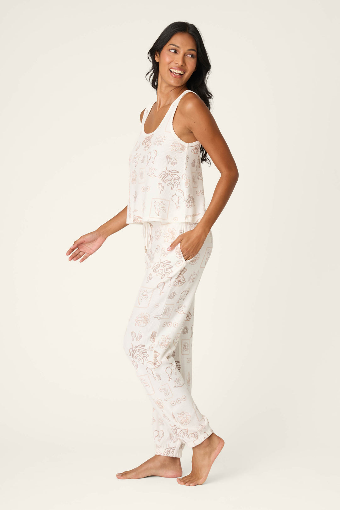 A woman with long dark hair smiles while posing barefoot in the Palms Away Bistro Botanicals Long PJ Set by P.J. Salvage, featuring a sleeveless printed top and matching pants, against a plain light background.