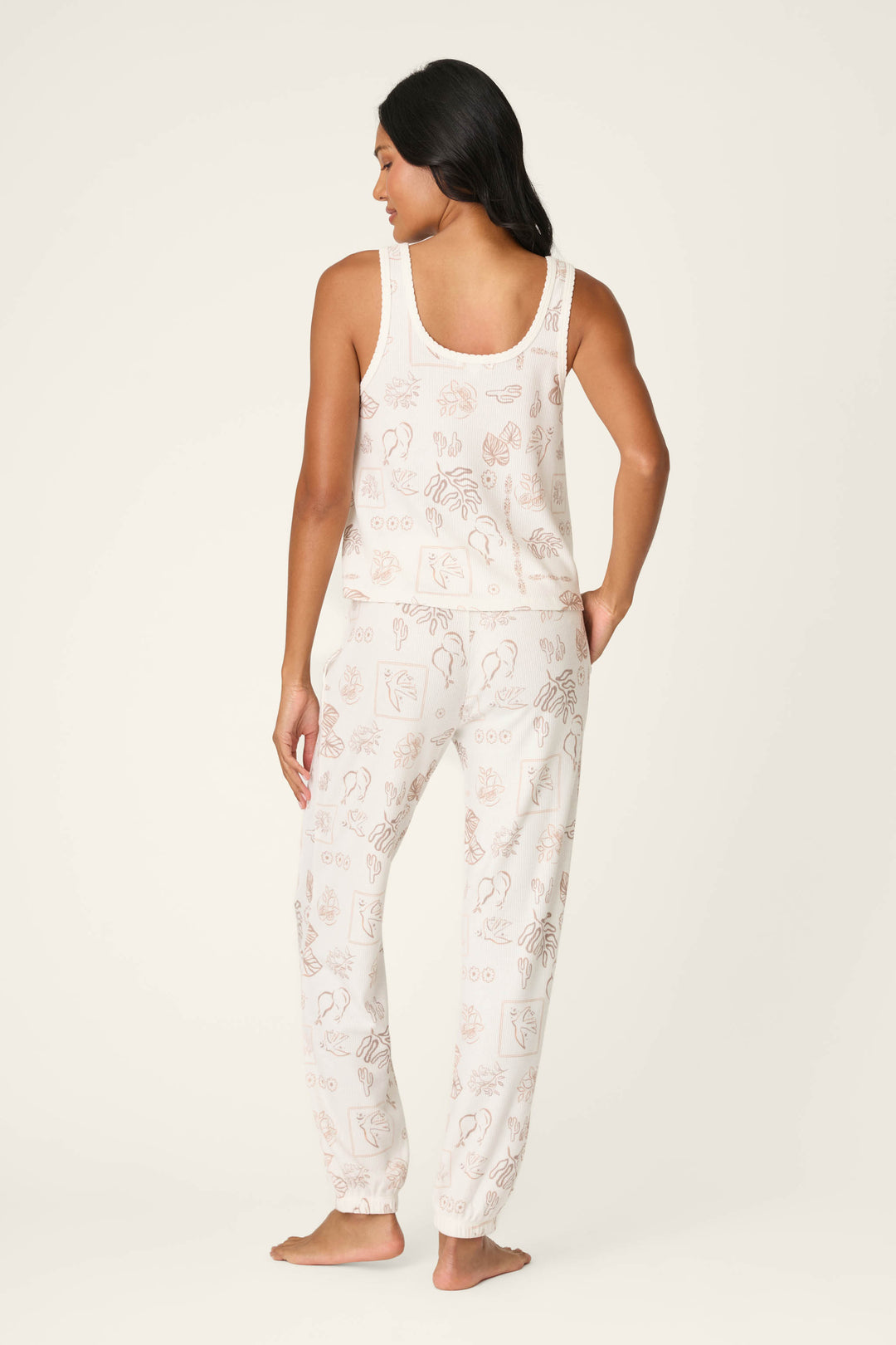 A woman with long dark hair stands barefoot, facing away, wearing the P.J. Salvage Palms Away Bistro Botanicals Long PJ Set—a sleeveless top and pants pajama set with a light cream background and subtle line art illustrations.