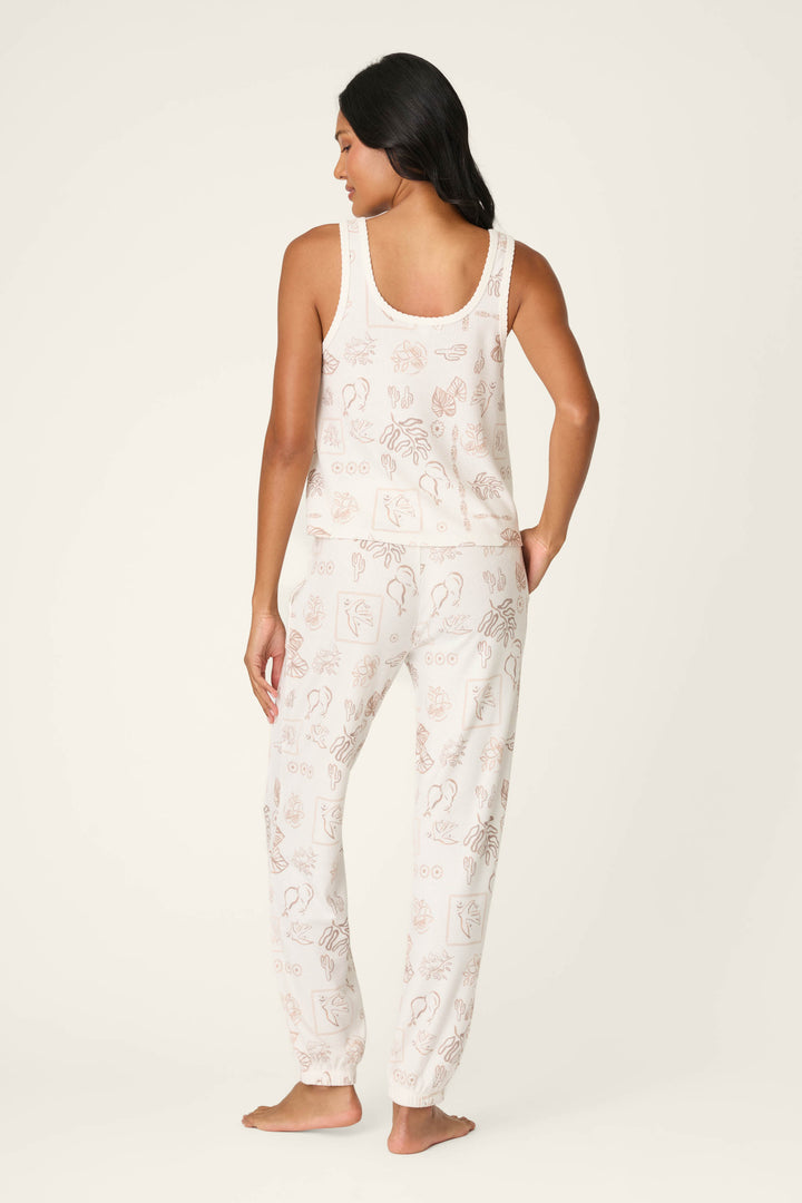 A woman with long dark hair stands barefoot, facing away, wearing the P.J. Salvage Palms Away Bistro Botanicals Long PJ Set—a sleeveless top and pants pajama set with a light cream background and subtle line art illustrations.