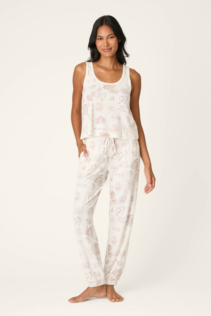 A woman stands barefoot against a plain background, smiling softly with one hand in her pocket. She wears the P.J. Salvage Palms Away Bistro Botanicals Tank Top, styled with relaxed-fit pajama pants.