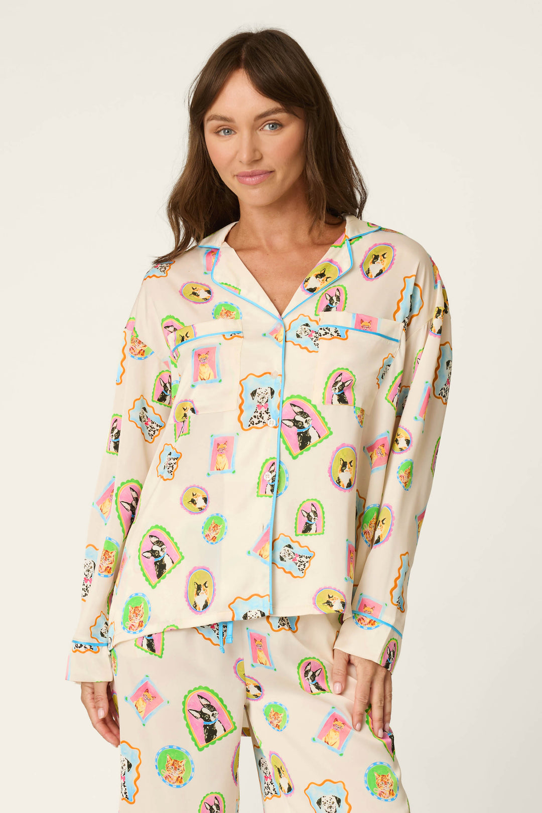 A woman wears the P.J. Salvage Picture Purrfect Pet Portrait Print Sateen Long Sleeve Top, featuring colorful cartoon animal faces, as she stands against a plain light background, smiling softly with one hand relaxed at her side.