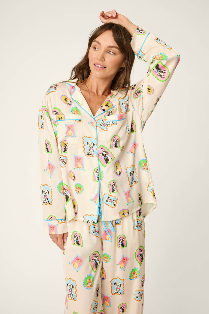 A woman wearing the P.J. Salvage Picture Purrfect Pet Portrait Print Sateen Long Sleeve Top—a cream-colored pajama top featuring colorful cartoon animal faces and blue trim—stands with one arm raised, gazing to the side with a gentle smile.