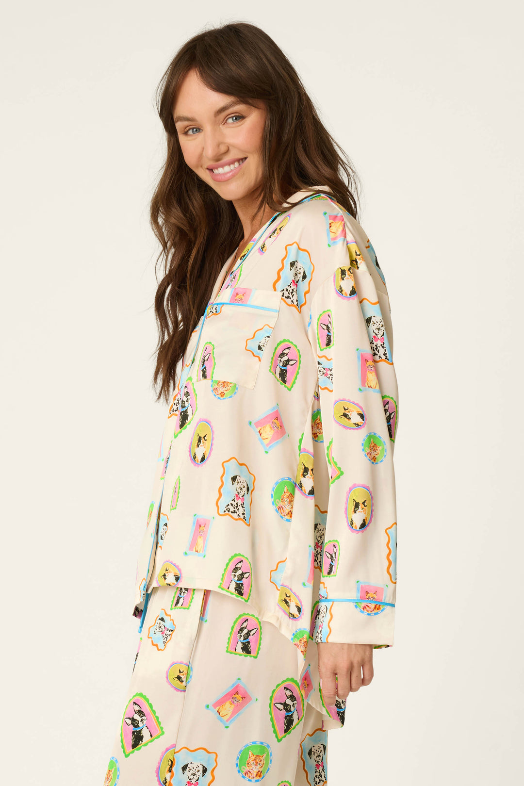 A woman with long brown hair smiles while wearing the P.J. Salvage Picture Purrfect Pet Portrait Print Sateen Long Sleeve Top, featuring cream-colored fabric adorned with colorful cartoon dog portraits, standing against a plain light background.