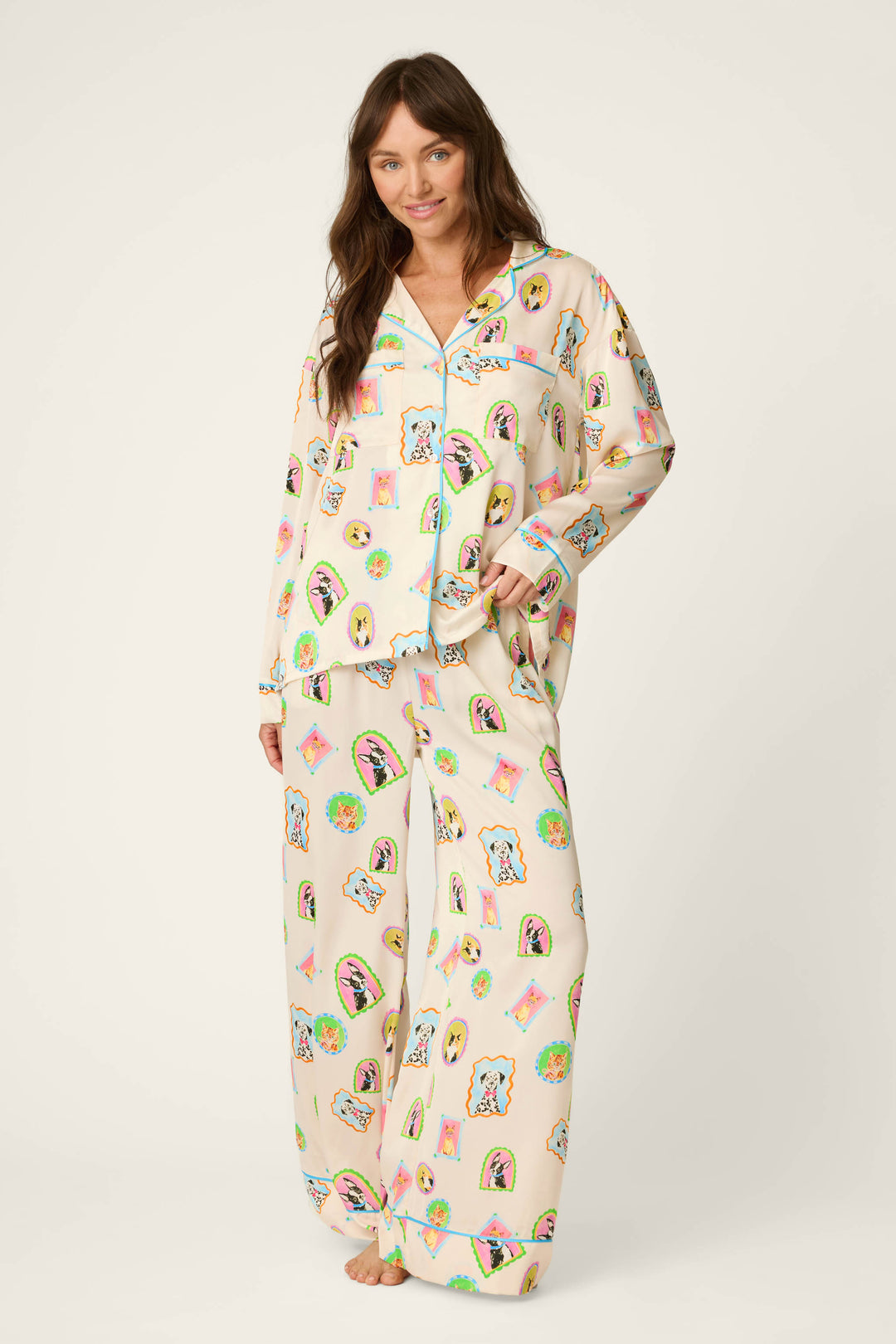 A woman with long brown hair wears the P.J. Salvage Picture Purrfect Pet Portrait Print Sateen Long Sleeve Top—loose, cream-colored pajamas adorned with colorful dog patterns. She stands barefoot on a white background, smiling with one hand in her pocket.