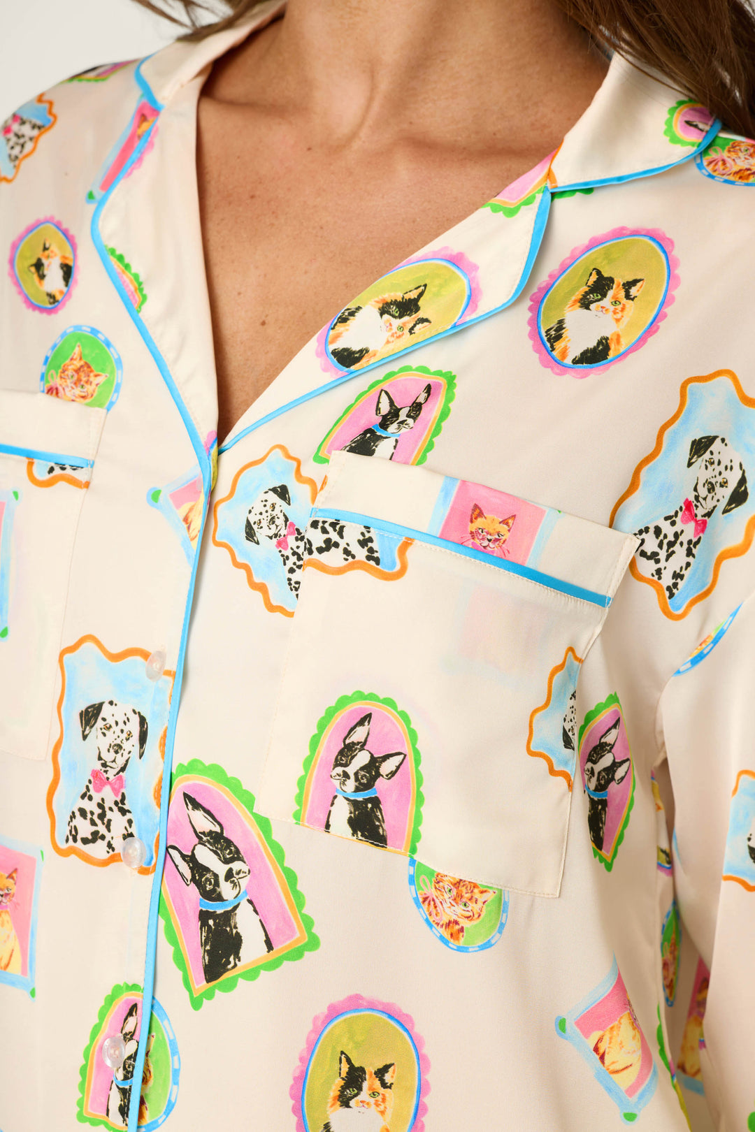 A woman wears the P.J. Salvage Picture Purrfect Pet Portrait Print Sateen Long Sleeve Top, featuring a cream-colored base with colorful framed dog and cat illustrations, a collar, blue piping, and a chest pocket.