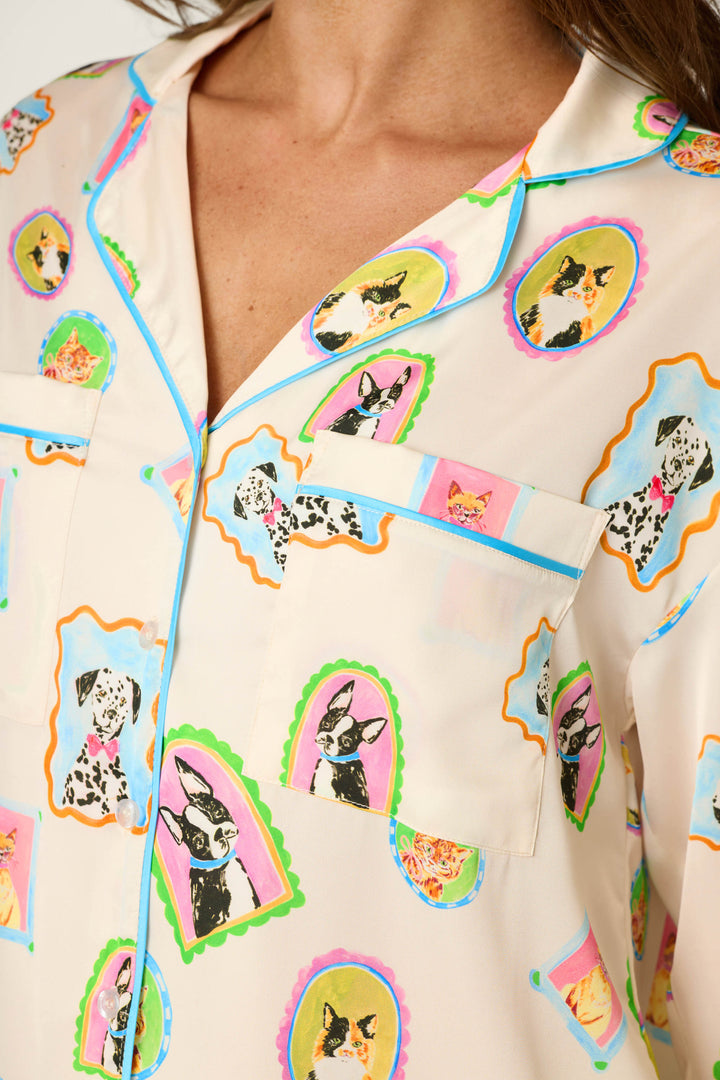 A woman wears the P.J. Salvage Picture Purrfect Pet Portrait Print Sateen Long Sleeve Top, featuring a cream-colored base with colorful framed dog and cat illustrations, a collar, blue piping, and a chest pocket.
