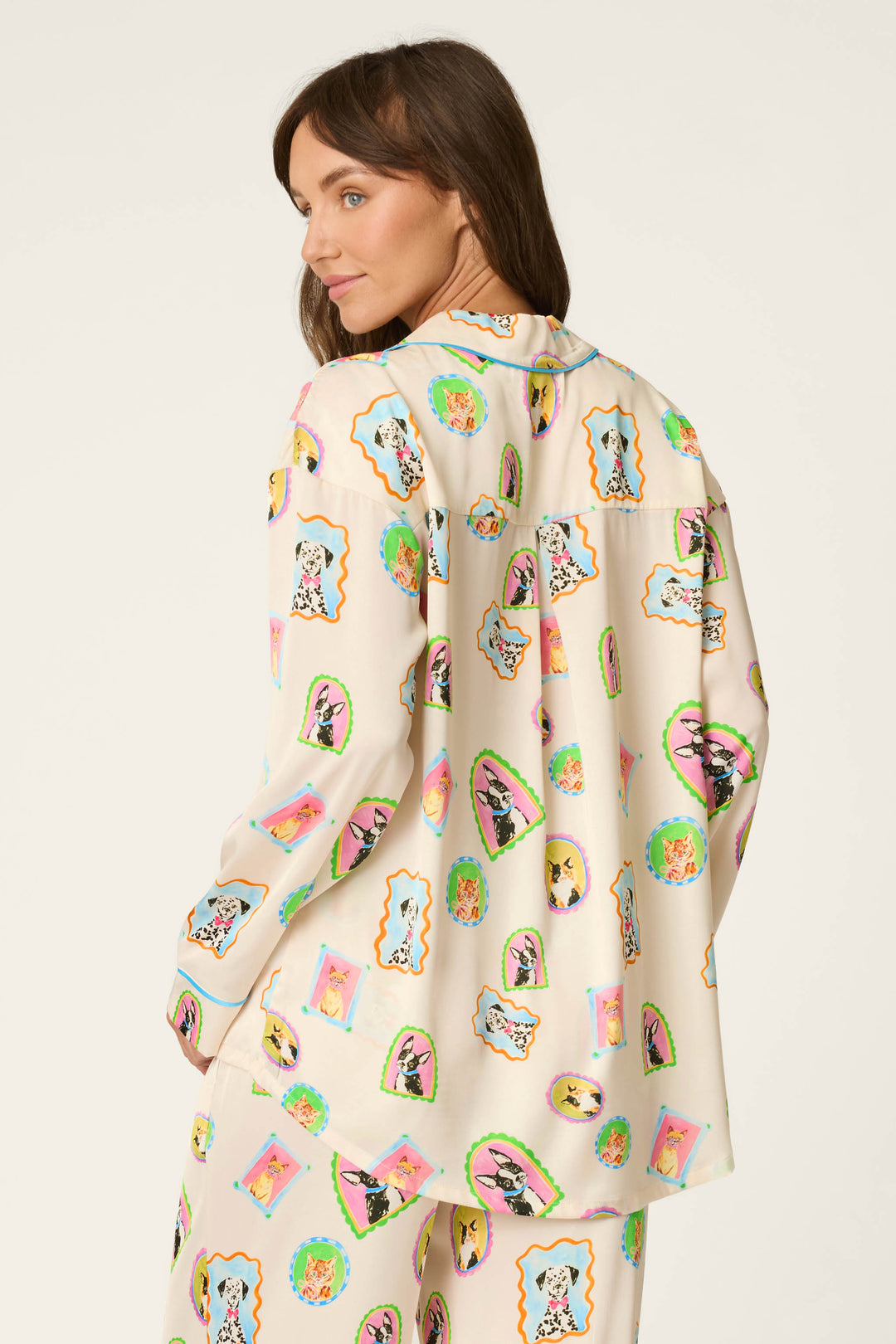 A woman models the P.J. Salvage Picture Purrfect Pet Portrait Print Sateen Long Sleeve Top, featuring cream fabric adorned with colorful, whimsical animal illustrations. She is shown from the back, glancing over her shoulder against a plain light background.