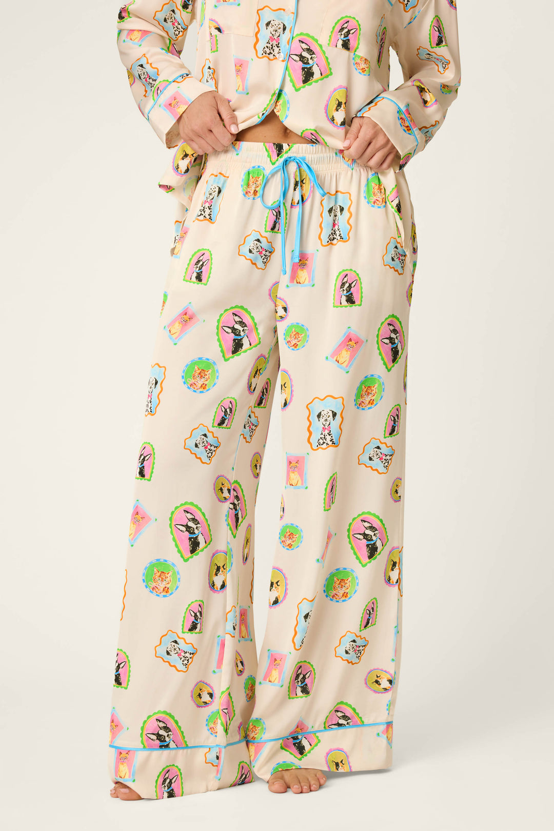 Model showcases the P.J. Salvage Picture Purrfect Pet Portraits Satin Pajama Pants, featuring a cream base adorned with vibrant cartoon dog faces and finished with a blue drawstring, standing barefoot against a light background. Only the lower half of the body is visible.