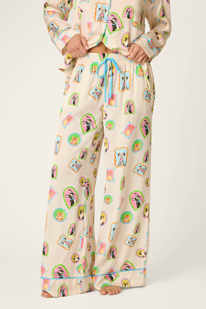 Model showcases the P.J. Salvage Picture Purrfect Pet Portraits Satin Pajama Pants, featuring a cream base adorned with vibrant cartoon dog faces and finished with a blue drawstring, standing barefoot against a light background. Only the lower half of the body is visible.