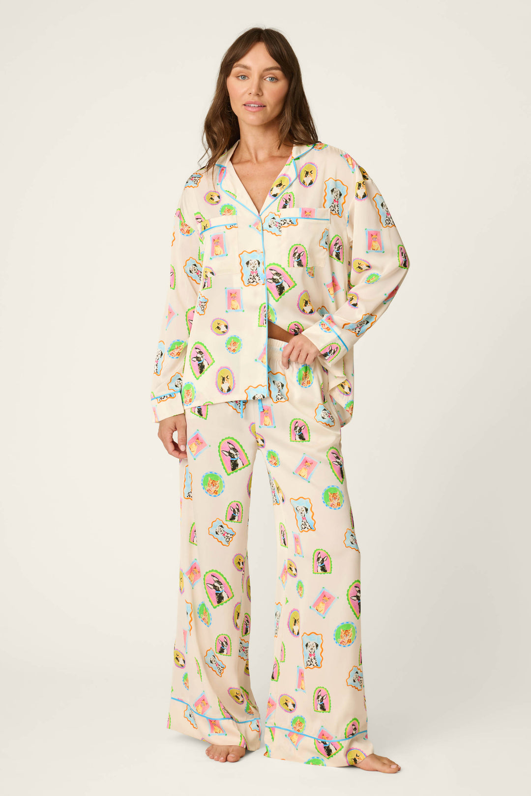A woman stands barefoot in P.J. Salvage's Picture Purrfect Pet Portraits Satin Pajama Pants, featuring loose, light-colored fabric adorned with playful, colorful cartoon pet faces and objects. She looks forward with a gentle smile against a plain background.