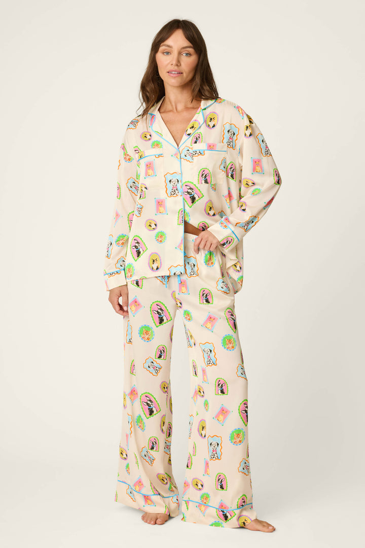 A woman stands barefoot in P.J. Salvage's Picture Purrfect Pet Portraits Satin Pajama Pants, featuring loose, light-colored fabric adorned with playful, colorful cartoon pet faces and objects. She looks forward with a gentle smile against a plain background.