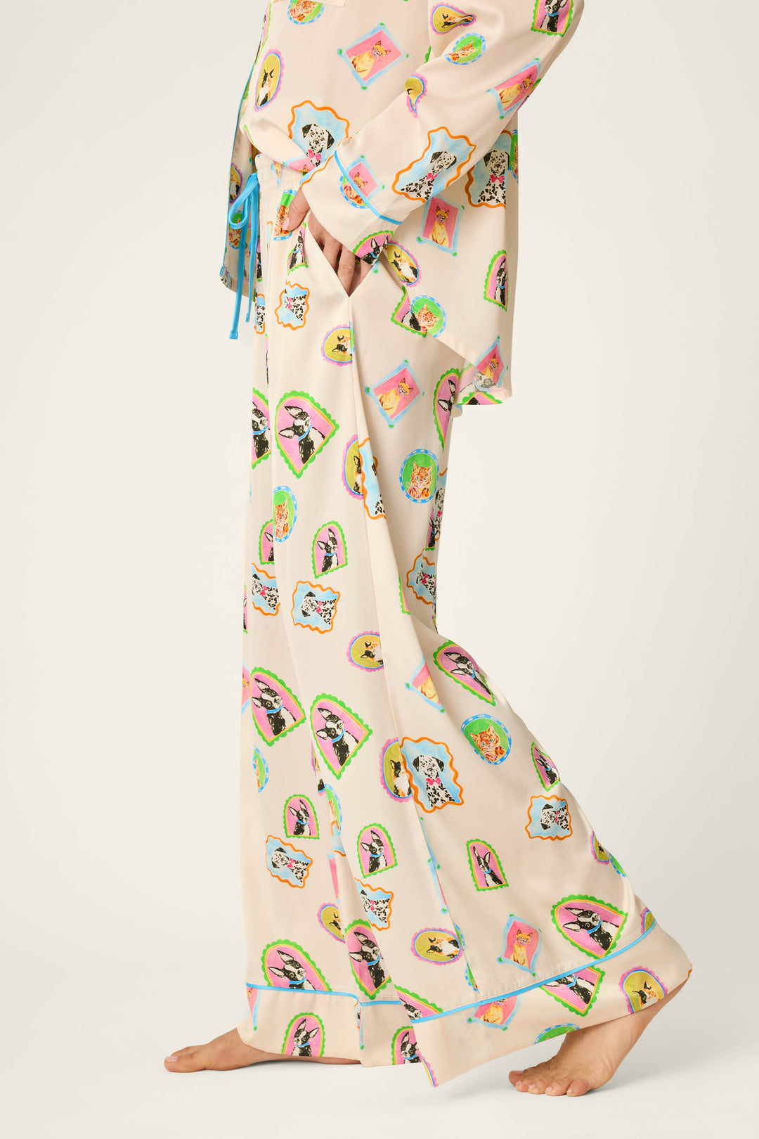 A person stands barefoot, partially visible from mid-torso down, wearing P.J. Salvage Picture Purrfect Pet Portraits Satin Pajama Pants in a loose, cream-colored style featuring colorful, cartoon-like animal faces.
