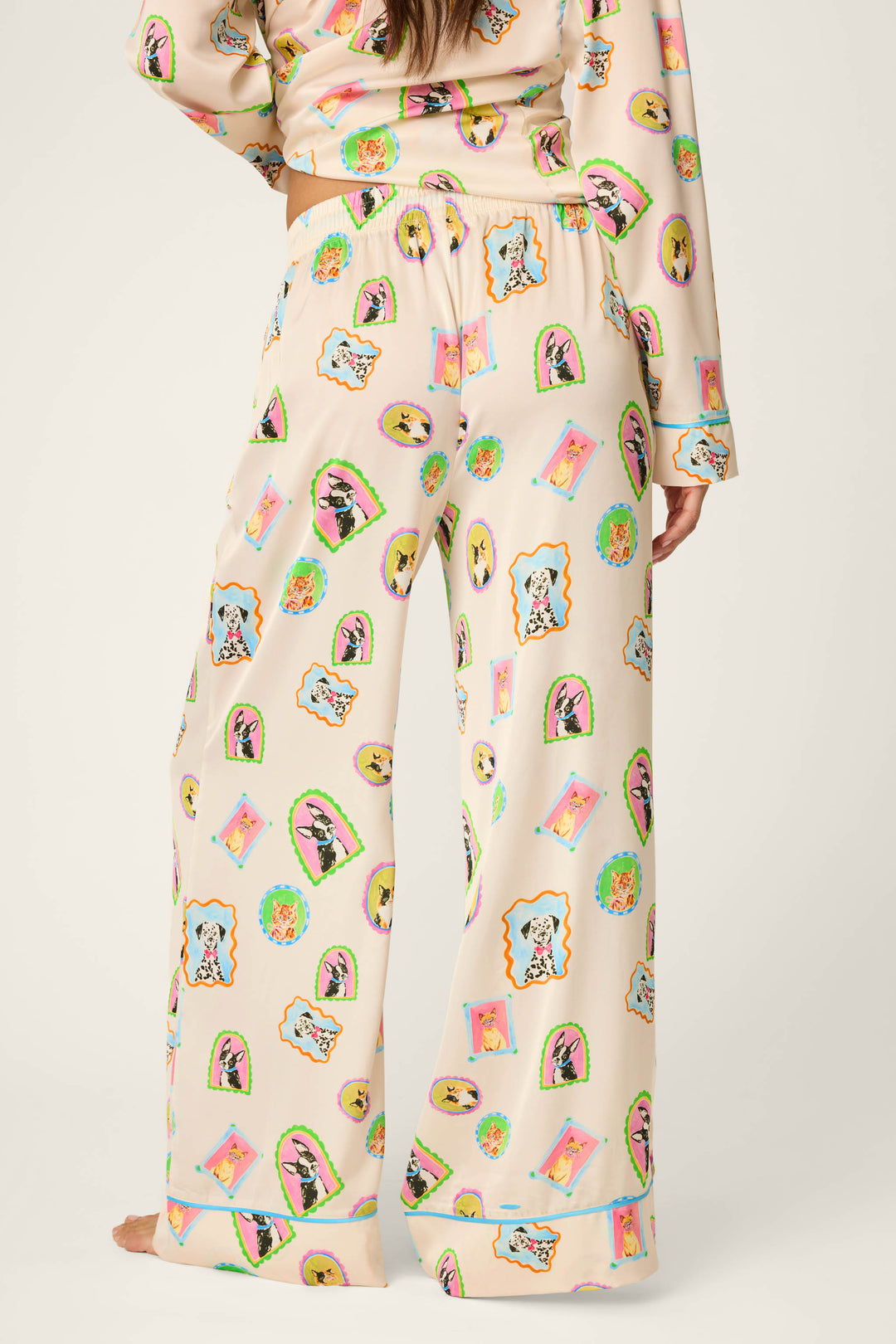 A person wearing P.J. Salvage Picture Purrfect Pet Portraits Satin Pajama Pants, featuring colorful cartoon dog and cat prints on light pink fabric, stands barefoot and faces away from the camera.