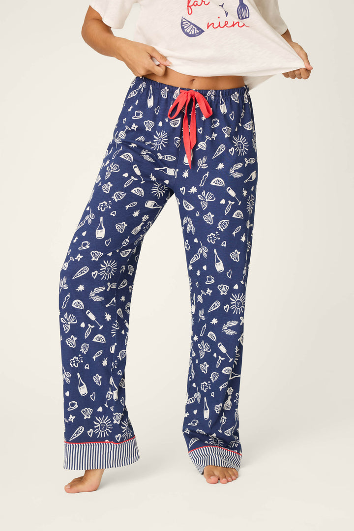 A person stands barefoot in P.J. Salvage Playful Prints Al Fresco Bistro Pajama Pants—blue with white playful prints and a red drawstring—paired with a white shirt, waistband revealed, and lower face not shown.