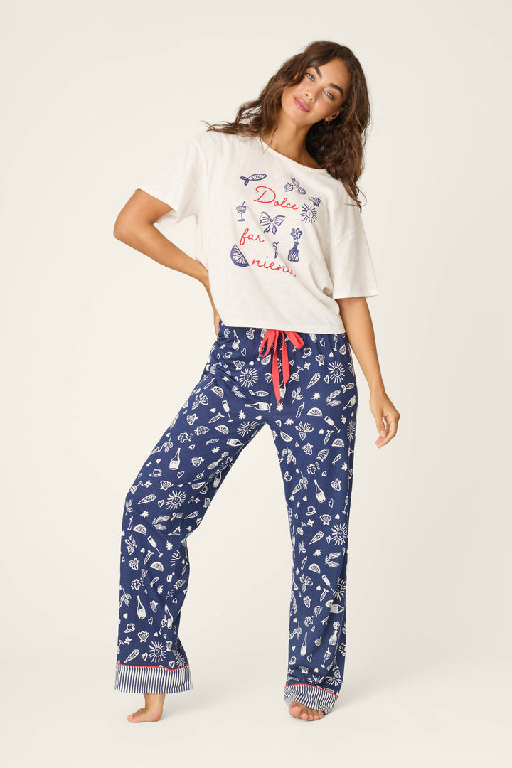 A woman poses against a plain light background wearing P.J. Salvage Playful Prints Al Fresco Bistro Pajama Pants—navy blue with a white seashell pattern and red drawstring—paired with a white T-shirt with red and blue graphics.