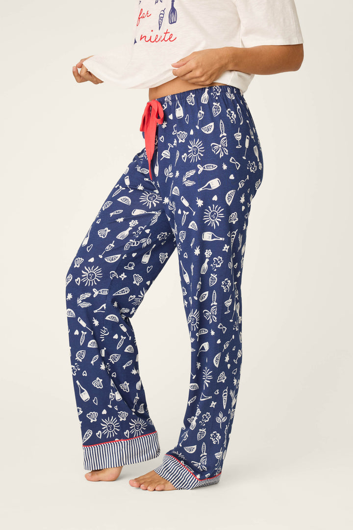 A barefoot person wears P.J. Salvage Playful Prints Al Fresco Bistro Pajama Pants with blue nautical and beach motifs, a red drawstring, and striped cuffs, lifting their white T-shirt to reveal the waistband.